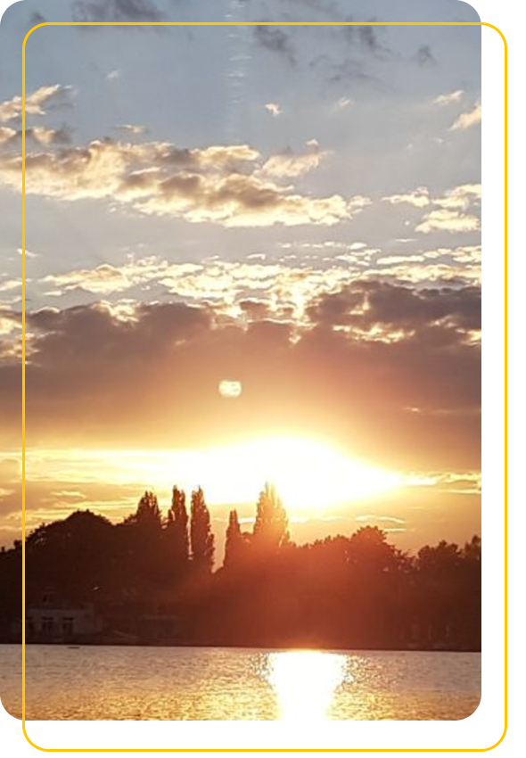 Bäume unter aufgehender/untergehender Sonne und vielen Wolken mit See im Vordergrund